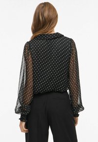 Zwarte blouse met witte polkadots, doorschijnende lange mouwen en een kraag. Heeft een losse pasvorm en elastische manchetten. Draagt bijbehorende zwarte broek.
