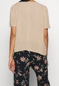 Beige Bluse mit kurzen Ärmeln und lockerem Schnitt; durchsichtiges Material, runder Ausschnitt und hinten etwas längerer Saum, kombiniert mit Hose mit Blumenmuster.