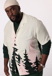 Korte mouwen shirt in crème met een groen en roze bosprint. Het shirt heeft een knoopsluiting aan de voorkant en een open kraag, gelaagd over een groene top.