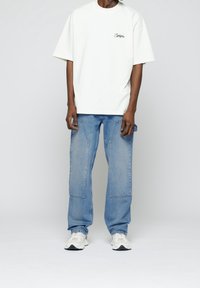 Camiseta blanca de gran tamaño con logo en el pecho, combinada con jeans sueltos de color azul claro con bolsillos de parche y zapatillas deportivas blancas.