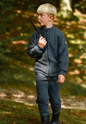 Grå uldjakke og bukser med lynlåslukning, struktureret stof og et lille logo-patch; barn holder en mørk fjer og bærer gummistøvler.