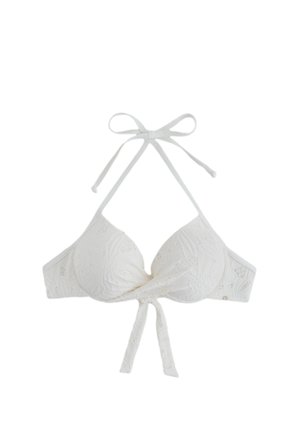 Femeie purtând bikini alb și ochelari de soare negri, stând desculță pe o plajă cu nisip, lângă o stâncă mare, ținând o geantă țesută de culoarea bronzului.