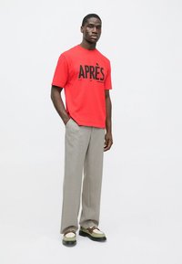 Κόκκινο βαμβακερό μπλουζάκι με μεγάλο μαύρο κείμενο «APRÈS.» Συνδυασμένο με ανοιχτό γκρι παντελόνια και λευκά, μπεζ και πράσινα αθλητικά παπούτσια με μαύρες σόλες.