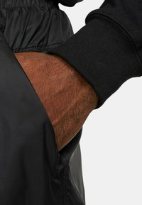 Svart ytterjacka med en slät, glänsande yta. Lätt elastiska ärmslut i svart, med en hand som sträcker sig in i en sidoficka.