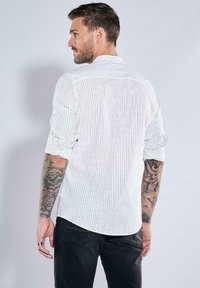 Weiß gestreiftes Hemd mit hochgerollten Ärmeln, leichtem Stoff, geschwungener Saum und detailliertem Tattoo am Arm. Getragen mit dunklen Hosen.