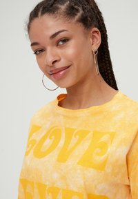 Żółta koszulka z kolorowym tie-dye z wyraźnym, wirującym napisem "LOVE" w kontrastującym odcieniu żółtego. Modelka nosi duże, okrągłe kolczyki.