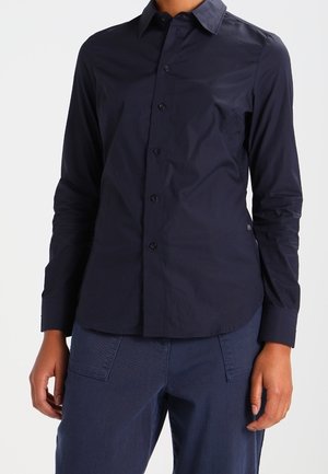 Person wearing a fitted, long-sleeve navy button-up shirt and dark blue pants, standing against a plain background.