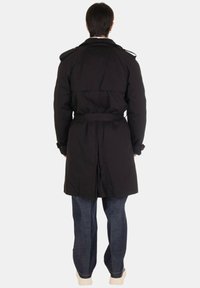 Cappotto trench nero con cintura, maniche lunghe, spalline e una apertura sul retro, abbinato a jeans blu e scarpe chiare.