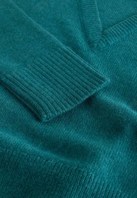 Pulover din lâna de culoare teal, cu un decolteu în V, având manșete și tiv cu nervuri. Materialul este moale, cu o textură netedă și variații subtile de culoare.