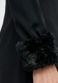 Μανίκι μαύρου παλτού με μαλακή, παχιά επένδυση από συνθετική γούνα που καλύπτει τον καρπό, πάνω σε απλό φόντο.