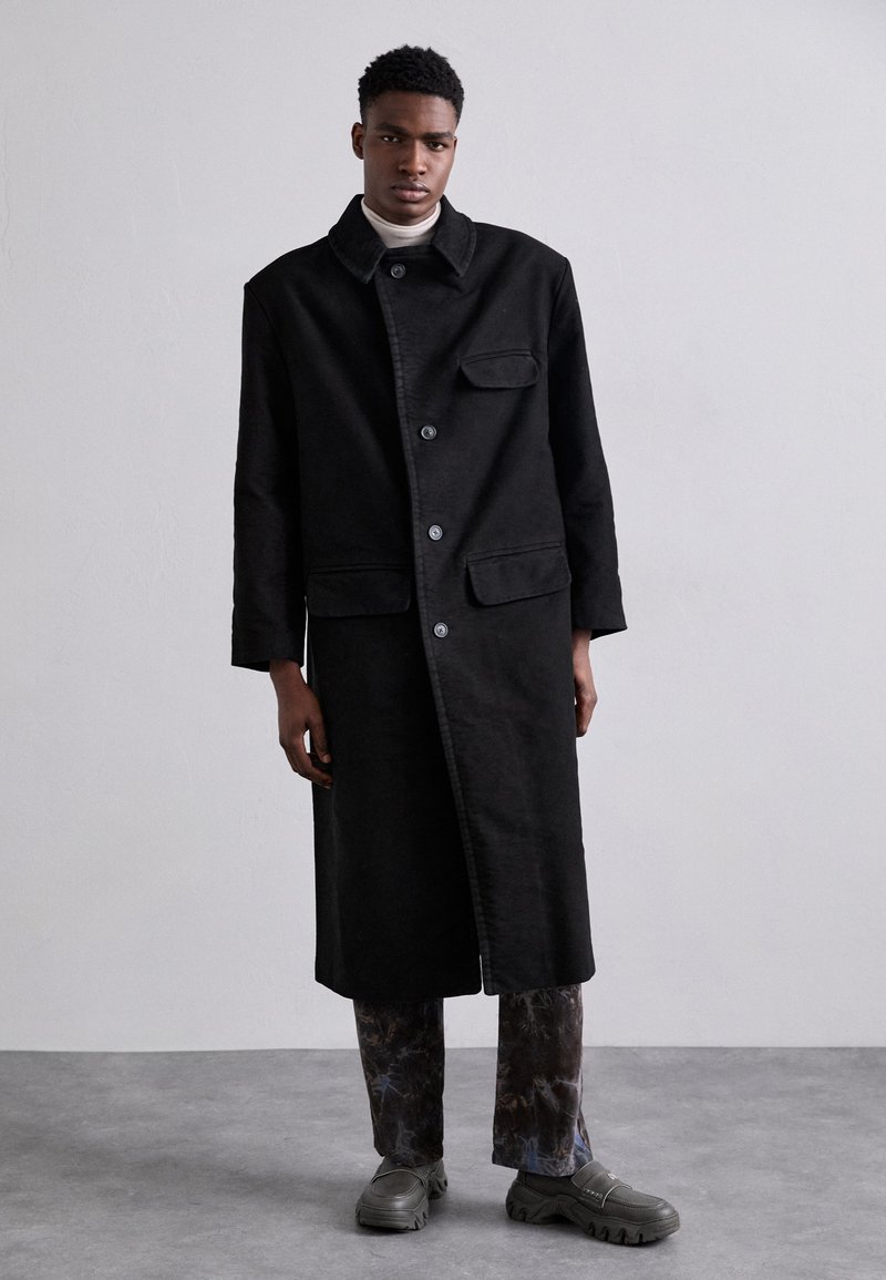 Black, long coat made of heavy fabric with a collar, front buttons, and side pockets, paired with patterned pants and gray footwear.