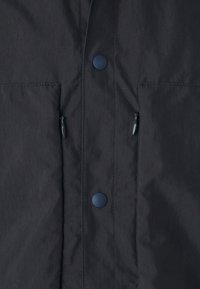 Dark navy jacket with a matte texture, featuring two zippered pockets, a row of snap buttons, and a streamlined design.