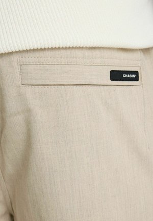 Beige broekzak met een zwart rechthoekig label met de tekst "CHASIN'", gedeeltelijk bedekt door een witte geribbelde trui.