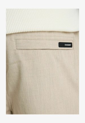 Beige broekzak met een zwart rechthoekig label met de tekst "CHASIN'", gedeeltelijk bedekt door een witte geribbelde trui.