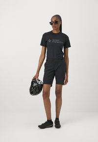 Svart sykkelantrekk med kortermet skjorte med logo og matchende shorts. Tilbehøret inkluderer svarte solbriller og en svart hjelm.