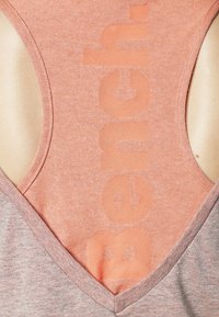 Top à manches sans épaules en pêche et gris avec une découpe en V dans le dos. Présente un grand texte "Bench" décoloré au centre. Tissu doux et texturé.