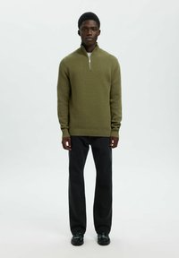 Alyvuogių žalios spalvos užtrauktuku užsisegantis megztinis tekstūrinio mezgimo, derinamas su juodomis tiesiomis kelnėmis ir juodais batais. Paprastas, minimalistinis dizainas.