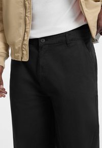 Black trousers made of a smooth fabric with a fitted design, featuring a button closure and side pockets. A beige bomber jacket is visible.
