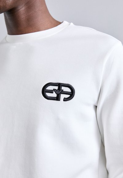 Sweatshirt branco com decote redondo e um logo bordado preto texturizado no peito, em fundo de tecido liso.