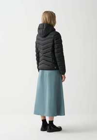 Zwarte, puffy, gewatteerde jas met chevronstiksels op de rug, gecombineerd met een lange, lichtblauwe rok en zwarte schoenen.