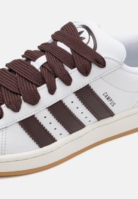 Zapatilla blanca de cuero con detalles en marrón y gruesos cordones marrones. Presenta tres rayas marrones y la palabra "CAMPUS" impresa en el lateral. Suela de goma texturizada.