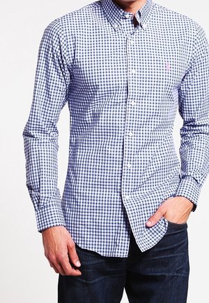 Homme portant une chemise à carreaux bleue et blanche avec une main dans la poche et un jean bleu foncé, sur un fond blanc.