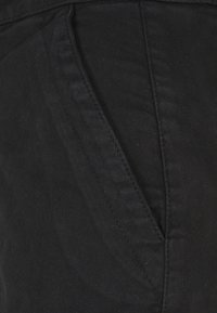 Detailní záběr černých kalhot z látky s viditelným diagonálním bočním kapesem a propracovaným šitím.