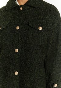 Giacca verde con texture realizzata in tessuto tessuto, dotata di colletto, bottoni dorati e due tasche frontali con chiusura a patta.