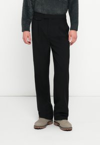 Μαύρο παντελόνι με εφαρμογή κατά παραγγελία, πιέτες και λεπτομέρεια με ένα κουμπί. Κατασκευασμένο από απαλό ύφασμα· συνδυασμένο με γκρι ανοιχτά μποτάκια αστραγάλου.