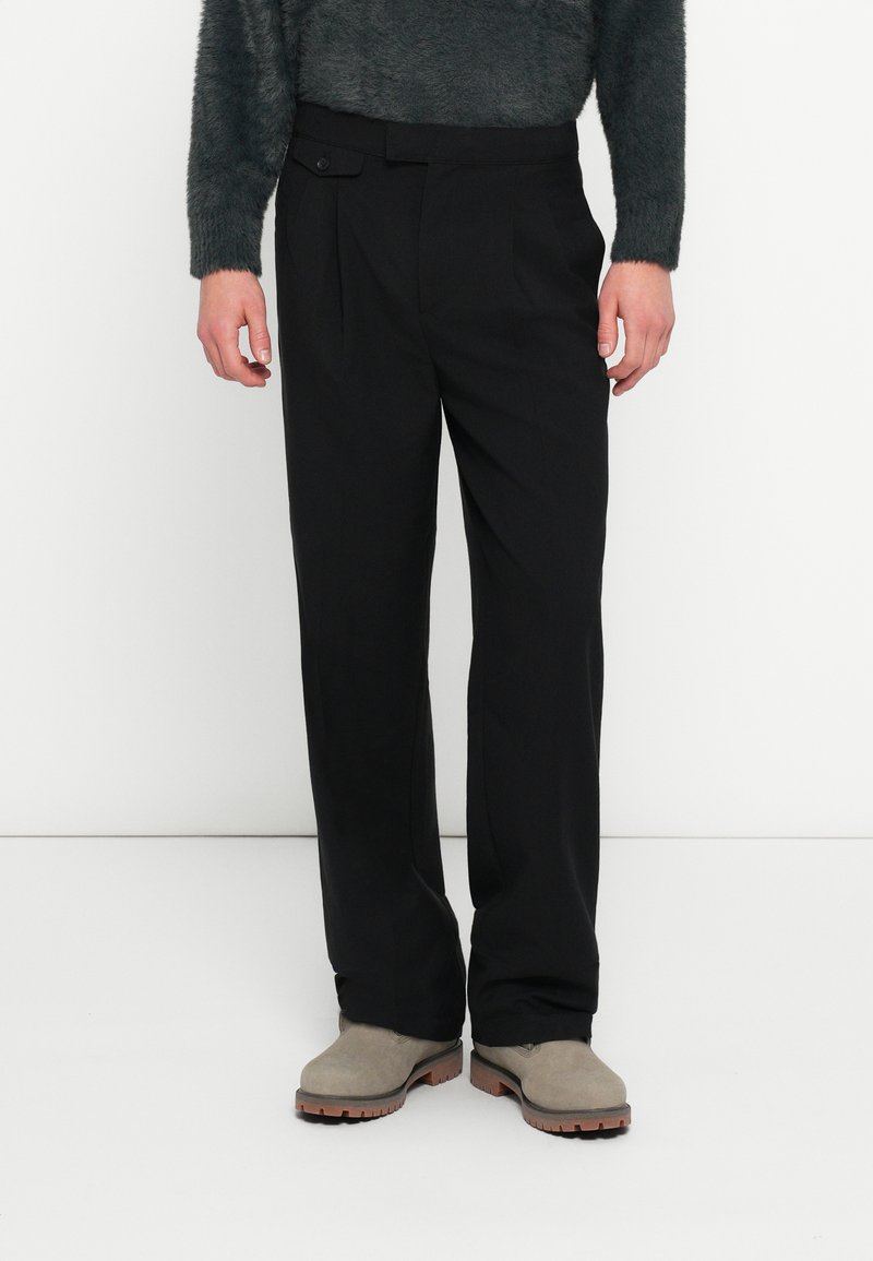 Μαύρο παντελόνι με εφαρμογή κατά παραγγελία, πιέτες και λεπτομέρεια με ένα κουμπί. Κατασκευασμένο από απαλό ύφασμα· συνδυασμένο με γκρι ανοιχτά μποτάκια αστραγάλου.