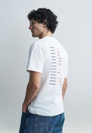 Görög hajú, szemüveges fiatal férfi oldalra fordulva, fehér mintás pólót és kék farmert visel, sima világos háttér előtt áll.