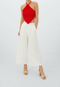 Haut licou rouge en tricot avec un design asymétrique, associé à un pantalon blanc à jambes larges. Des talons compensés tressés couleur tan complètent la tenue.