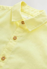 Camisa de lino amarilla con un collar clásico, dos botones de madera y una textura fina con sutiles detalles de costura.