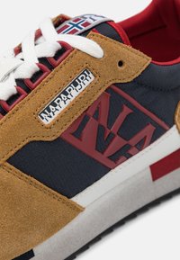 Sneaker con una combinazione di tessuto blu navy e suede marrone, dettagli rossi, lacci bianchi e un'etichetta del marchio visibile. Suola intermedia testurizzata.
