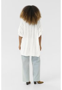 Vit stickad tunika med ett zigzag-mönster, löst sittande och med vida ärmar. Matchad med ljusblå jeans och bruna klackstövlar.