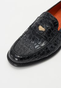 Zwarte loafers met een krokodillenstructuur, voorzien van een gouden accent aan de voorkant en een oranje binnenvoering. Platte zool en een afgeronde neus.