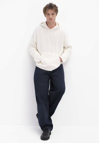 Cremefarbener Strick-Hoodie mit Fronttasche, kombiniert mit locker sitzenden dunkelblauen Hosen und schwarzen Schuhen, für ein lässiges, geschichtetes Outfit.