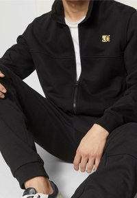 Chaqueta negra con cierre de cremallera y cuello alto, con un logo dorado, combinada con pantalones negros a juego y zapatos blancos. Tejido suave y texturizado.