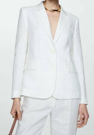 Frau trägt einen weißen, maßgeschneiderten Blazer und passende Hose, hält den Riemen einer braunen Handtasche und trägt eine goldene Choker-Halskette.