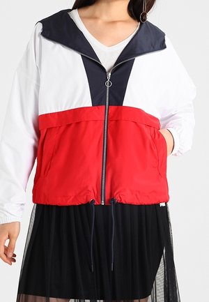 Une femme porte une veste à capuche zippée blanche, bleu marine et rouge avec une jupe noire, sur un fond uni.