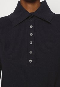 Marinblå virkad polo skjorta med bred krage, som har sex knappar framtill. Texturen är ribbad med en slät yta på knapparna.