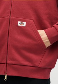 Κοντινό πλάνο του χεριού ενός ατόμου στην μπροστινή τσέπη ενός κόκκινου φούτερ με φερμουάρ Dickies, με ριμπ τελείωμα στη μέση και στις μανσέτες.