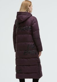 Purple puffer coat with a hood. Boxy design featuring stitched panels and black graphic text accents. Zippered side pockets.