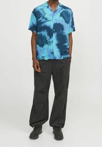 Hombre vistiendo una camisa de patrón en tonos azul y negro de manga corta, pantalones negros sueltos con puños elásticos y zapatillas negras contra un fondo liso.