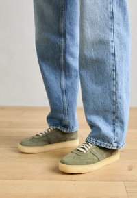 Green suede sneakers with beige rubber soles, featuring white laces and a textured design, paired with light blue jeans.