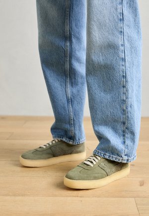 Baskets en daim vert avec semelles en caoutchouc beige, dotées de lacets blancs et d'un design texturé, associées à un jean bleu clair.