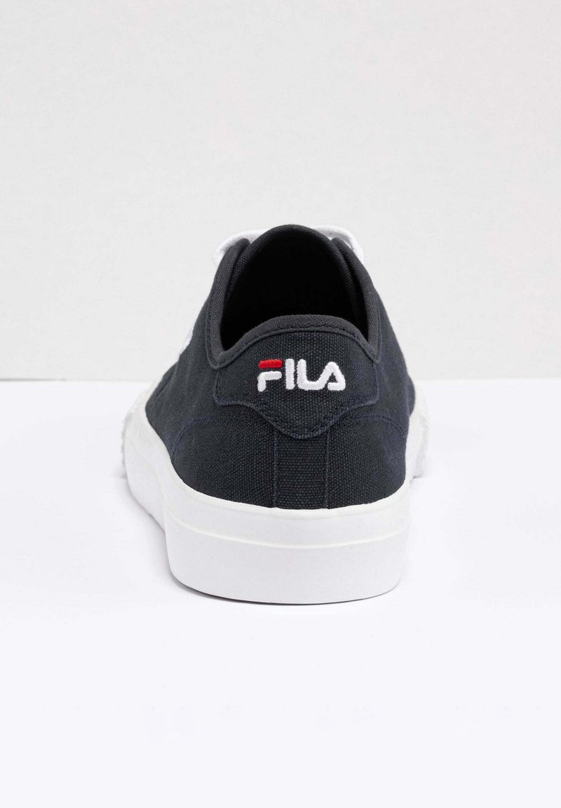 Zapatilla de lona gris oscuro con suela de goma blanca sólida, que presenta un parche de logo rojo y azul en la parte trasera. Tacón redondo y diseño simple.
