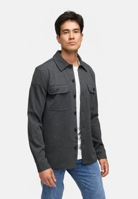 Grå skjorte med langeærmer, to brystlommer og en struktureret krave, båret over en hvid skjorte og parret med blå jeans.