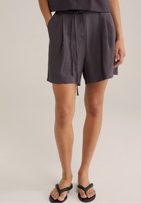 Šedé šortky vyrobené z lehkého materiálu, s volným střihem, stahovacími šňůrkami v pase a bočními kapsami, doplněné černými žabkami.