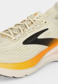 Gros plan d'une chaussure de sport beige et orange avec un swoosh noir, des perforations détaillées et des lacets noués sur fond blanc.